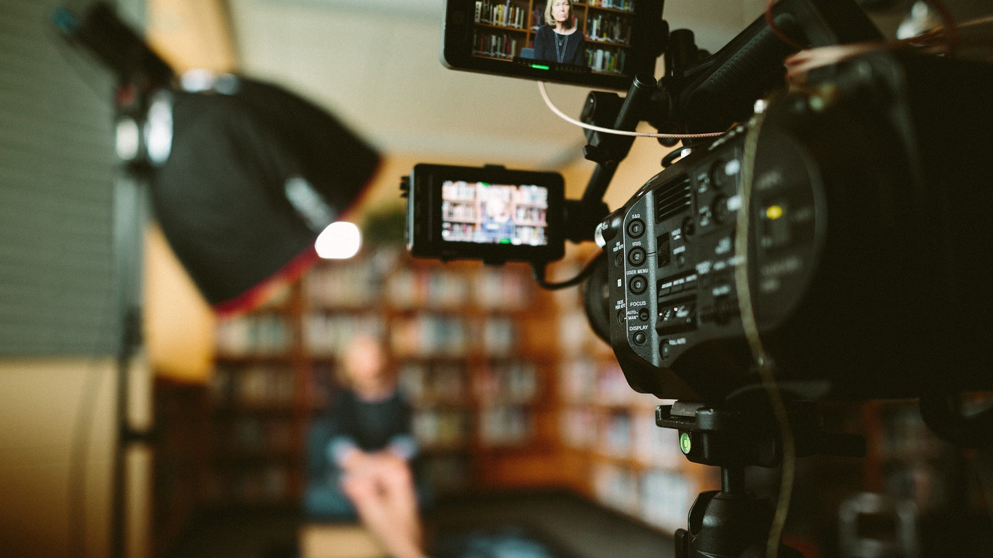 An older professional woman being filmed in their home in front of a wall of books. The client is blurred and the filming equipment is in focus in right foreground of the shot.