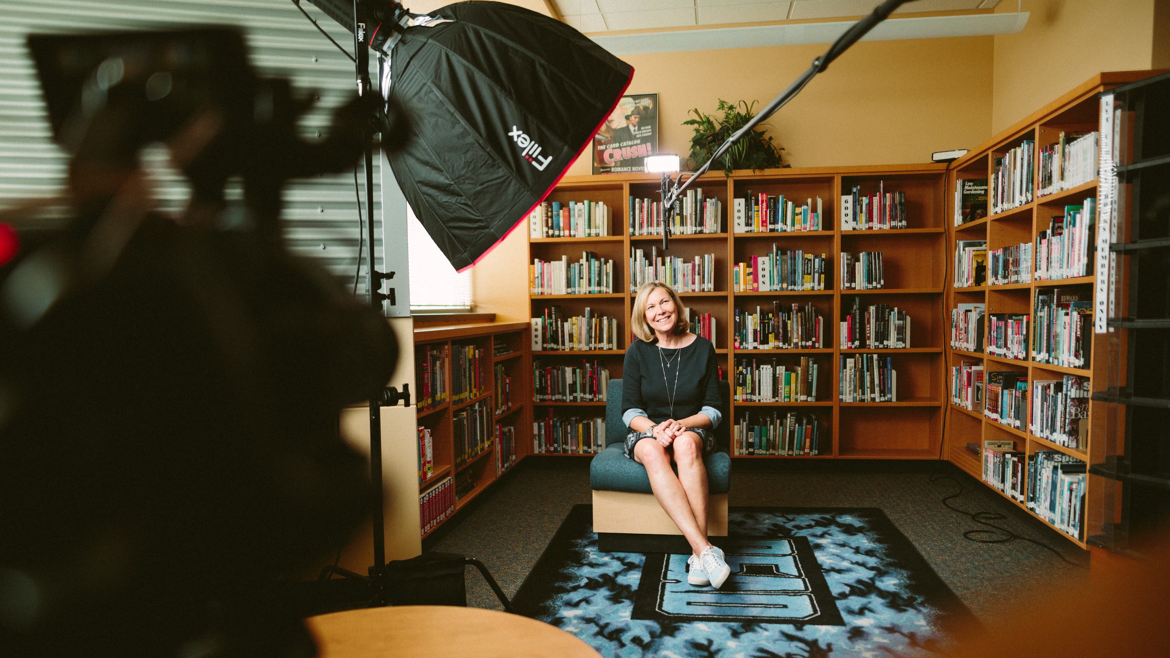 A woman being filmed in front of a wall of books smiling at the camera. Lighting and camera visible in frame on left of the image.
